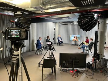 Studio mit Podiumsrunde aus vier Personen und einem zugeschalteten Gast auf einem Bildschirm, davor Kameras auf Stativen und eine Softbox
